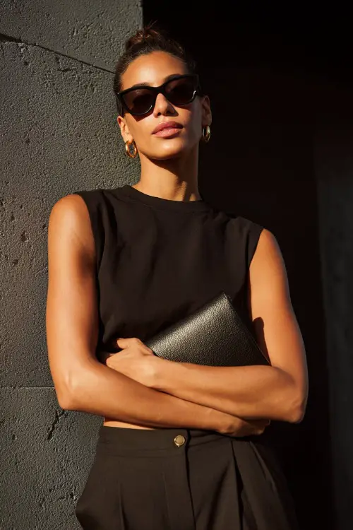 A woman wearing a monochrome black sleeveless top paired with tailored black trousers, styled with oversized sunglasses, a leather clutch, and minimalist gold hoops