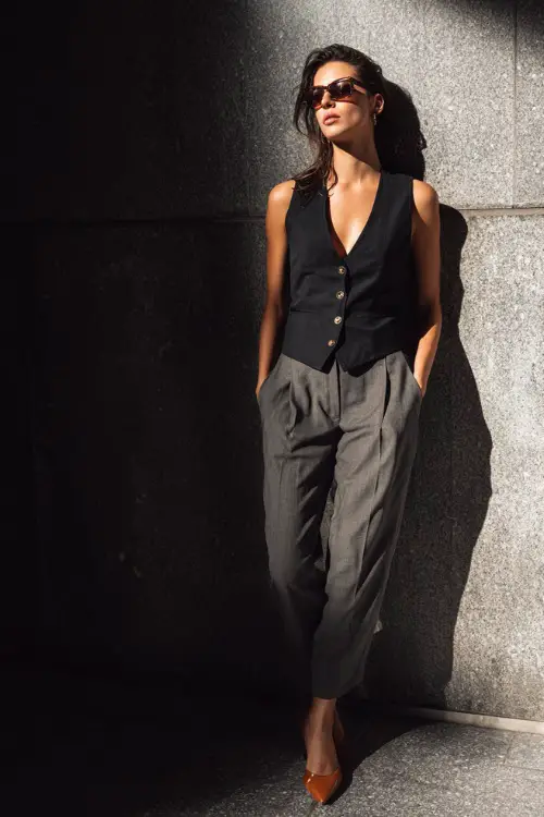 A woman wearing a structured sleeveless black vest worn as a top with low-rise tailored gray trousers, styled with pointed leather mules and slim rectangular sunglasses 