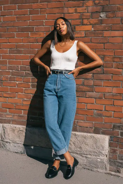 A woman wearing a white ribbed tank top tucked into high-waisted straight-leg blue jeans, paired with black loafers and a slim belt