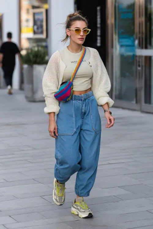 confident woman wearing a baby tee layered with a lightweight cardigan and oversized baggy jeans with chunky sneakers, accessorized with a colorful shoulder bag and tinted sunglasses