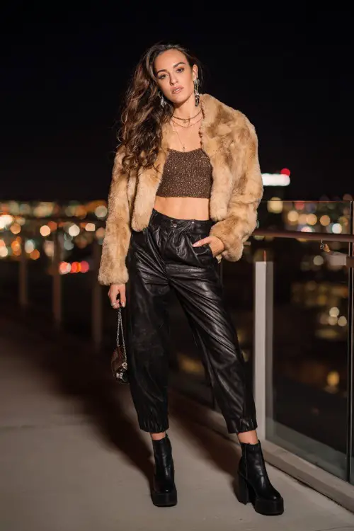 confident woman wearing a cropped faux fur jacket over a glittery crop top with low-rise leather pants and platform boots, accessorized with statement earrings and a mini bag