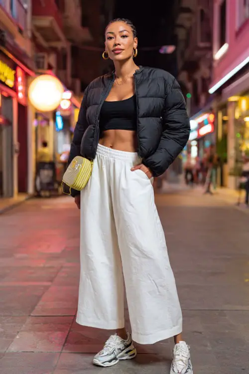 confident woman wearing a cropped puffer jacket over a tank top with low-rise wide-leg pants and chunky sneakers, accessorized with hoop earrings and a mini handbag 