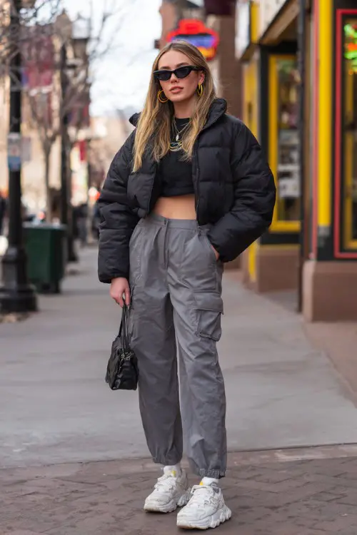 confident woman wearing a cropped puffer jacket with a baby tee and low-rise cargo pants with platform sneakers, accessorized with hoop earrings and a mini handbag 