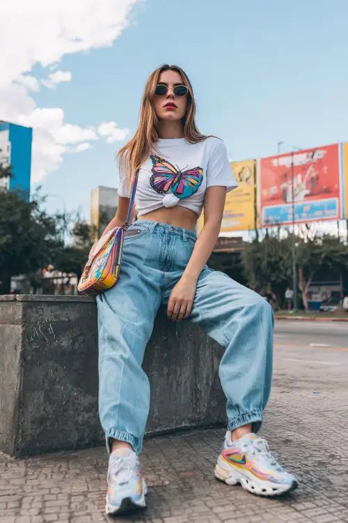 fashionable woman wearing a baby tee with a butterfly graphic and low-rise baggy jeans, chunky sneakers and a colorful shoulder bag, Y2K aesthetic outfit