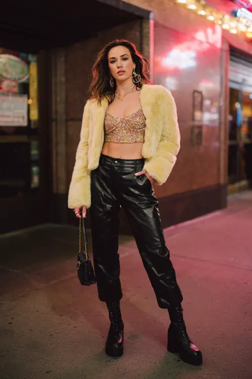 fashionable woman wearing a cropped faux fur jacket over a glittery crop top with low-rise leather pants and platform boots, accessorized with statement earrings and a mini bag