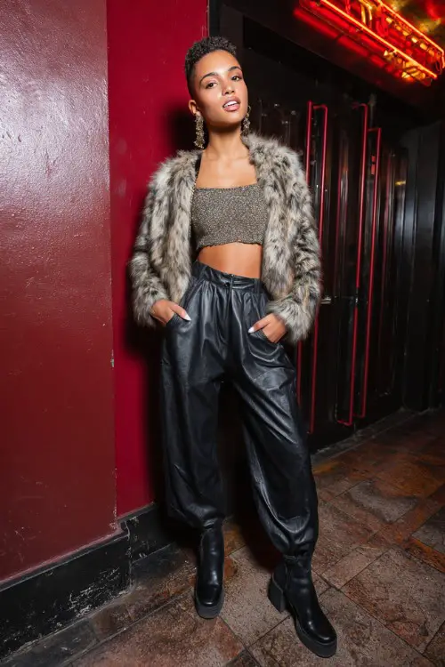 fashionable woman wearing a cropped faux fur jacket over a glittery crop top with low-rise leather pants and platform boots, accessorized with statement earrings and a mini bag