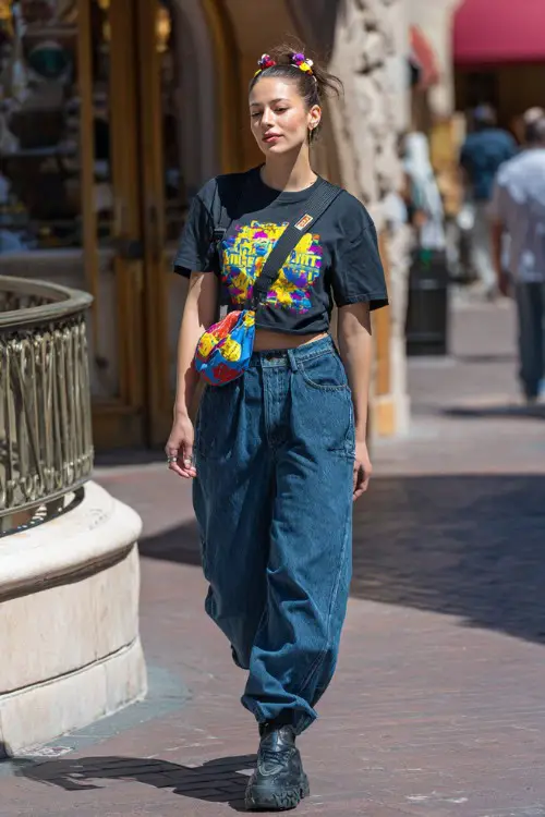fashionable woman wearing a graphic baby tee tucked slightly into oversized baggy jeans with platform sneakers, accessorized with a colorful shoulder bag and hair clips