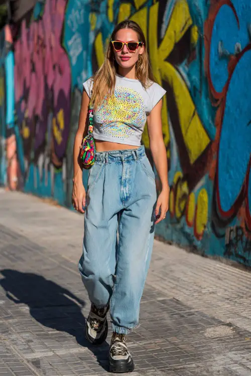 fashionable woman wearing a rhinestone baby tee with low-rise baggy jeans and chunky platform sneakers, accessorized with a colorful shoulder bag and tinted sunglasses