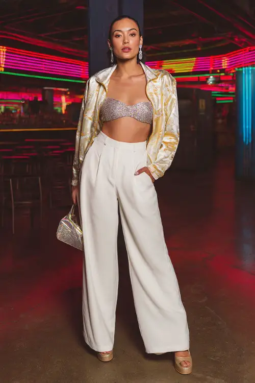 stylish woman wearing a shiny satin jacket over a glittery tube top with low-rise wide-leg pants and platform heels, accessorized with statement earrings and a mini bag