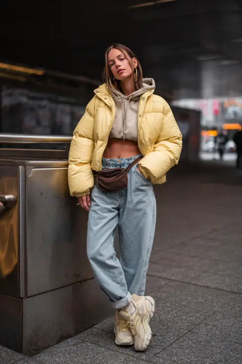 trendy woman wearing a puffer jacket over a cropped hoodie with baggy jeans and chunky sneakers, accessorized with hoop earrings and a shoulder bag, casual Y2K streetwear outfit with jacket