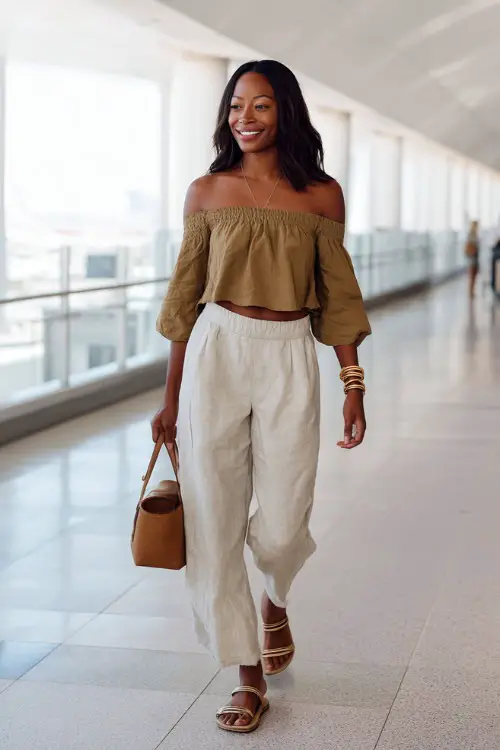 A Black woman wears a stylish off-shoulder top with relaxed linen pants, paired with slide sandals and layered gold accessories