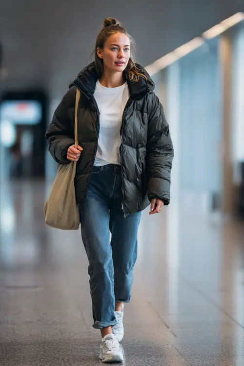 A woman wears a puffer jacket layered over a simple t-shirt and straight-leg jeans, paired with sneakers and a tote bag
