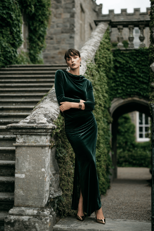 Elegant modest evening outfit with a dark green velvet maxi dress, gold heels, and statement jewelry beside a historic stone staircase