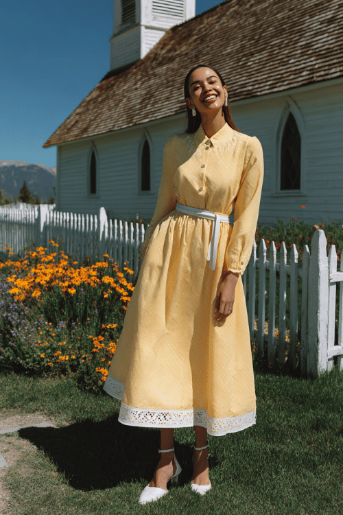 Modest church outfit with a yellow long sleeve midi dress, white belt, and white flats beside a charming countryside chapel and flower garden