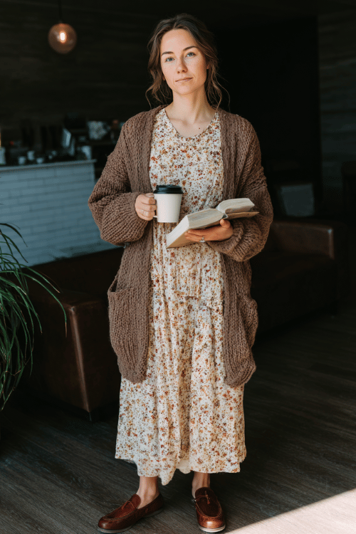 Young Christian woman in a cozy modest casual outfit, oversized knit cardigan layered over a floral ankle-length dress, brown leather flats, holding a Bible and coffee cup 