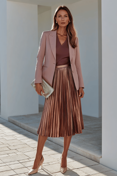 a chic woman in her 40s wearing a pleated midi skirt in dusty rose paired with a fitted long-sleeve top and a tailored blazer, holding a clutch bag