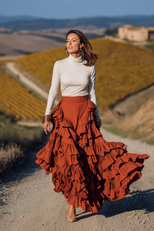 a striking woman in her 40s wearing a dramatic tiered ruffle maxi skirt in terracotta paired with a fitted long-sleeve mock-neck top in ivory