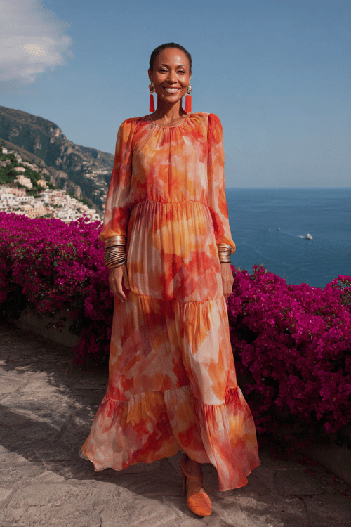 woman in her 50s wearing a gorgeous long-sleeve tiered maxi dress in warm sunset watercolor print with flowing layers of chiffon in shades of peach, tangerine and blush