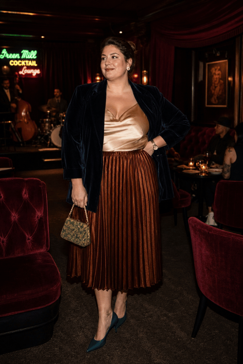 Plus-size woman in a navy velvet blazer, champagne satin camisole, and bronze pleated midi skirt.