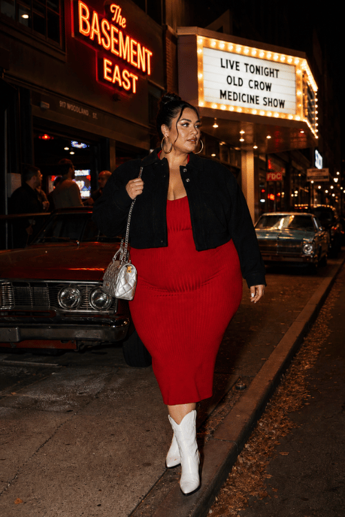 Plus-size woman in a red ribbed midi dress with a cropped black jacket and white cowboy boots.