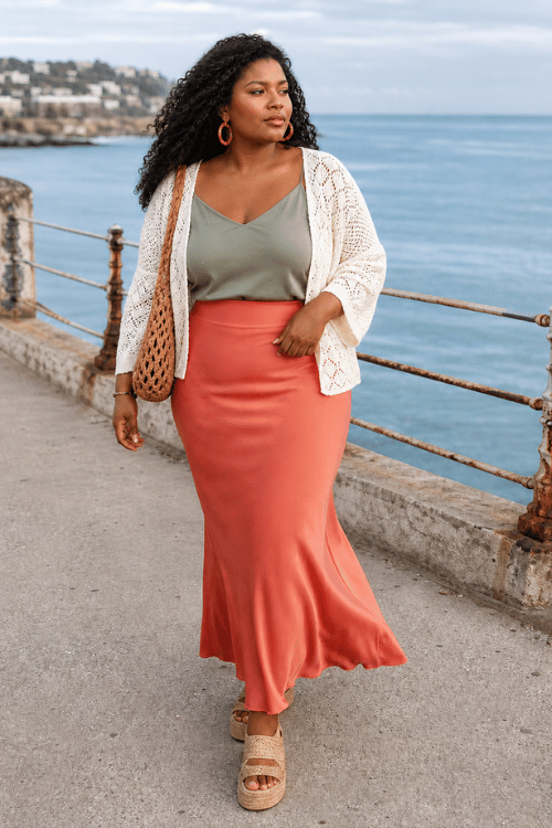Plus-size woman in a sage camisole, coral satin maxi skirt, and white crochet cardigan.