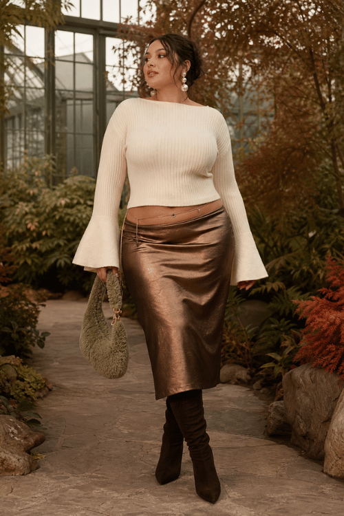 Plus-size woman wearing a cream ribbed sweater with flared sleeves and a bronze metallic midi skirt with dark brown knee-high boots and a textured shoulder bag.