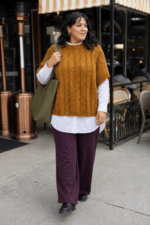 Plus-size woman wearing a mustard cable knit sweater vest over a white button-down shirt with plum wide-leg trousers and black ankle boots.