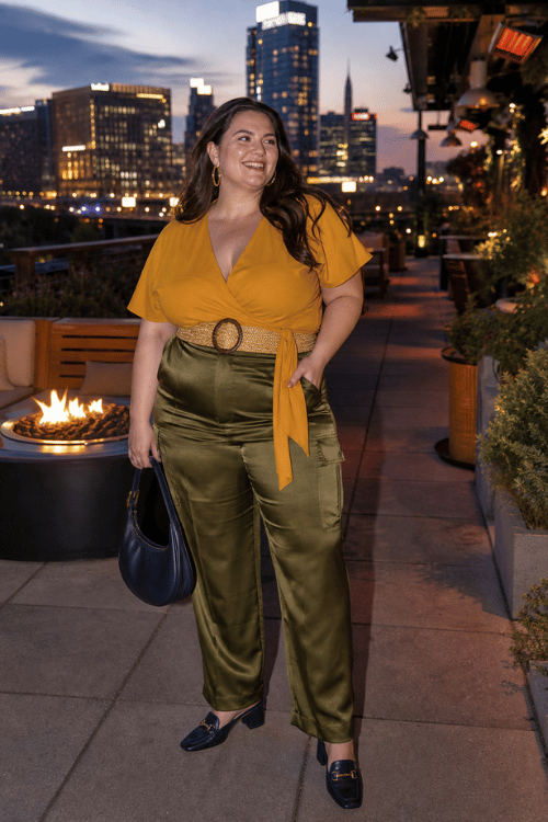 Plus-size woman wearing a mustard wrap blouse, olive satin trousers, and black loafers on a Nashville rooftop.