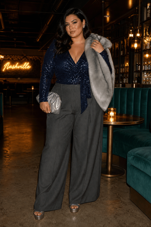 Plus-size woman wearing a navy sequin wrap top with gray wide-leg trousers and silver platform sandals.