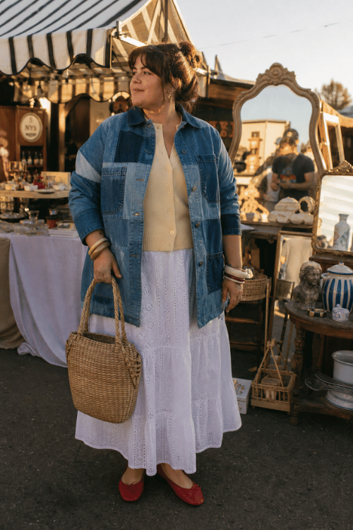 Plus-size woman wearing a patchwork denim jacket over a cream knit cardigan with a white eyelet maxi skirt, red ballet flats, stacked bangles, and a woven straw tote at an outdoor vintage market.
