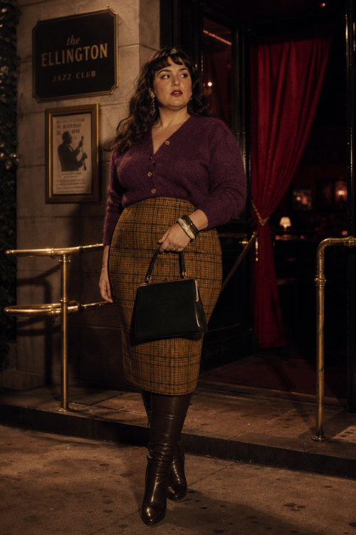 Plus-size woman wearing a plum cardigan with a mustard plaid pencil skirt, tall brown leather boots, stacked bracelets, and a structured black handbag outside a jazz club.