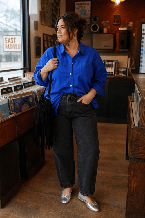 Plus-size woman wearing a royal blue button-up shirt, black jeans, and silver ballet flats inside a record store.