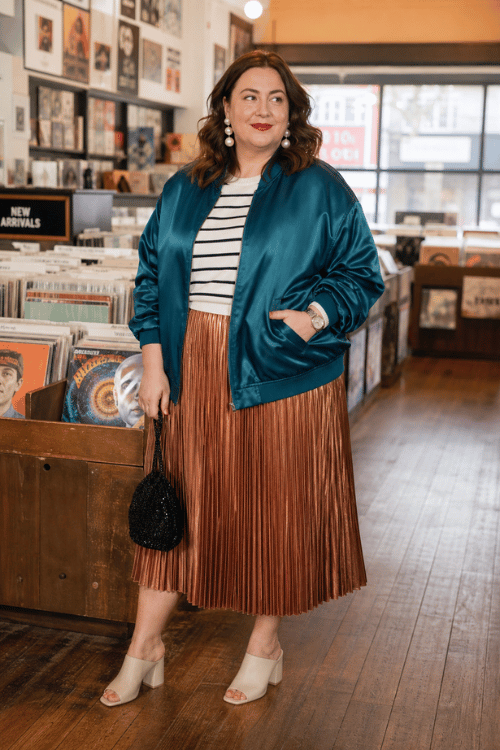 Plus-size woman wearing a teal satin bomber jacket with a striped top and a bronze pleated midi skirt.