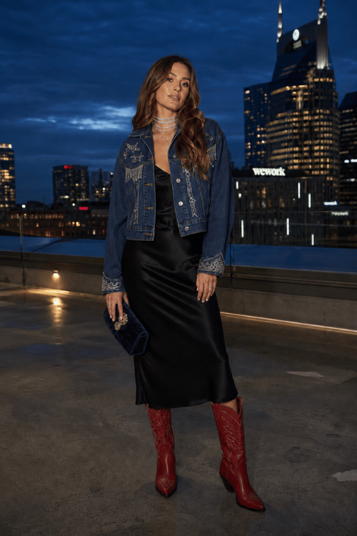 Woman in a black satin slip dress layered with a denim jacket and red cowboy boots.