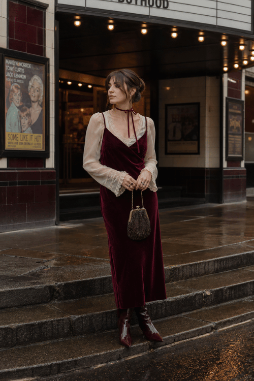 Woman in a burgundy velvet slip dress layered over a sheer blouse with burgundy ankle boots.