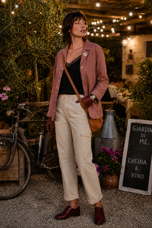 Woman in a dusty rose utility jacket, black lace camisole, cream pants, and burgundy loafers at a cozy outdoor restaurant.