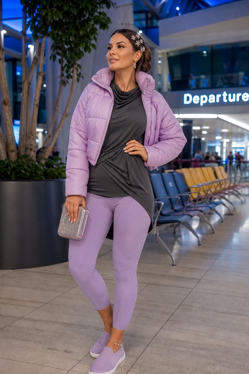 Woman in a lavender puffer jacket, gray draped top, lavender leggings, and slip-on sneakers holding a glitter clutch at an airport.