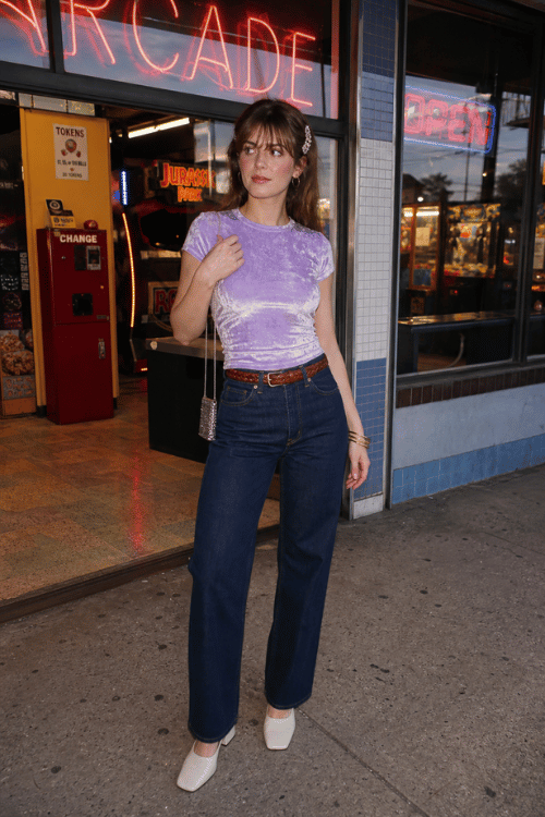 Woman in a lavender velvet top with dark straight-leg jeans and white pointed heels.