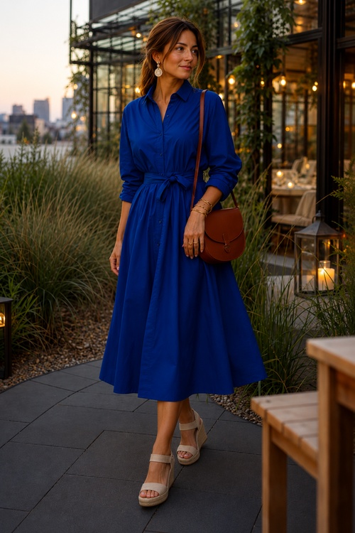 Woman in a royal blue belted shirt dress with beige wedge sandals and a brown crossbody bag at an outdoor restaurant.
