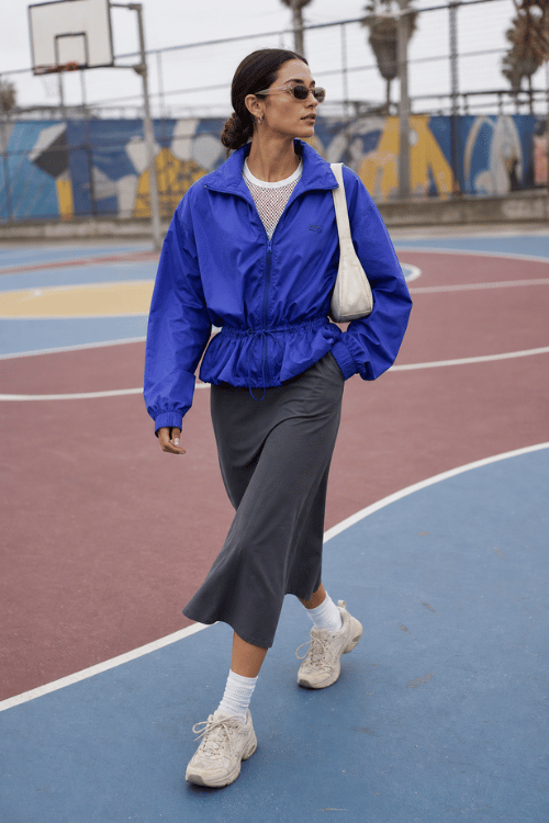 Woman in a royal blue windbreaker with a gray midi skirt and white sneakers.