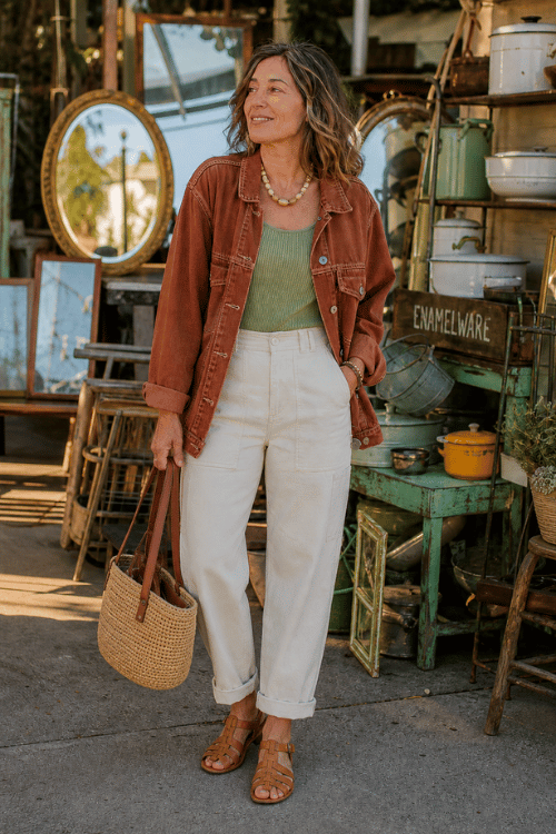 Woman in a rust denim jacket with a sage tank top, white jeans, and brown sandals.