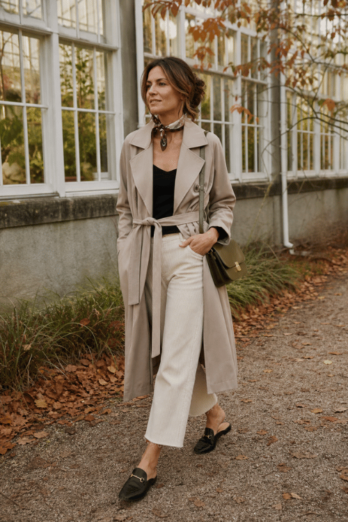 Woman wearing a beige belted trench coat over a black top with ivory cropped trousers and black loafers.