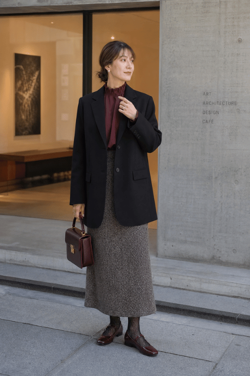 Woman wearing a black blazer over a burgundy blouse with a brown tweed midi skirt, burgundy Mary Jane flats, and a structured handbag.
