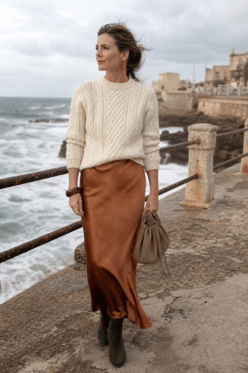 Woman wearing a cream cable knit sweater with a copper satin midi skirt and ankle boots.
