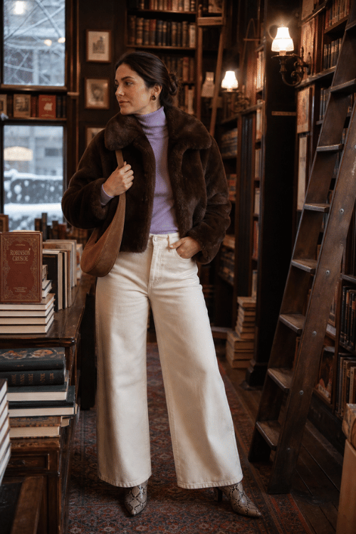 Woman wearing a dark brown faux fur jacket over a lavender turtleneck with white wide-leg jeans, snakeskin ankle boots, and a tan leather shoulder bag in a cozy bookstore.