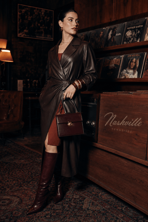 Woman wearing a dark brown leather wrap coat over a rust knit dress with knee-high leather boots in a Nashville record store.
