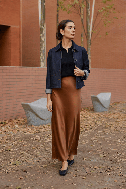Woman wearing a dark denim jacket over a black knit top with a bronze satin maxi skirt and navy ballet flats.