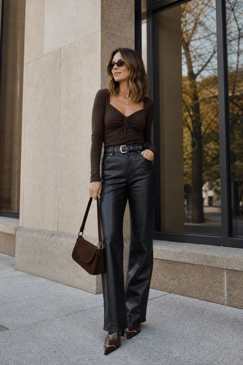 Woman wearing a fitted chocolate brown top with black leather trousers, pointed-toe heels, and a brown shoulder bag.