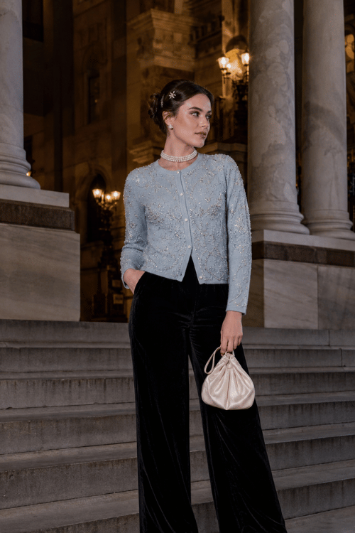 Woman wearing a light blue embellished cardigan with black velvet wide-leg trousers, white heeled sandals, a pearl choker, crystal hair clips, and a cream satin pouch for an elegant evening.