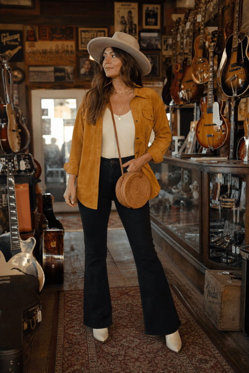 Woman wearing a mustard overshirt with black flare jeans, a cowboy hat, and white boots.
